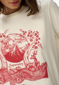 Kremowy t-shirt z czerwonym nadrukiem przedstawiającym górę, słońce, drzewa i jelenia. Tekst „Superdry” wkomponowany w ilustrację.