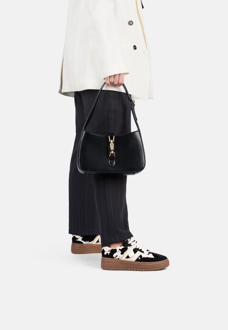Borsa a tracolla in pelle nera con hardware dorato, silhouette curvata, portata da una persona in giacca bianca e pantaloni a righe, abbinata a sneakers con fantasia.
