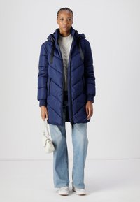 Doudoune marine avec fermeture éclair à l'avant, matelassage en chevrons et capuche. Associée à un jean large bleu clair et des baskets blanches, tenant un petit sac.