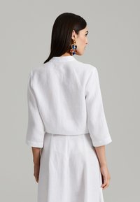 Bela lanena srajca z visokim ovratnikom in rokavi do tri četrtine, ki ima teksturirano površino in sproščen kroj. Zadnji pogled prikazuje preprost dizajn.