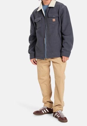 Person trägt eine marineblaue Fleecejacke mit weißem Kragen, hellblaues Hemd, beigefarbene Hose und braune Sneaker mit weißen Streifen.