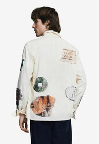 Kreemika värvi pikkade varrukatega särk, millel on suured, värvikad graafilised trükised artefaktidest ja tekstuuridest seljal ja varrukatel.