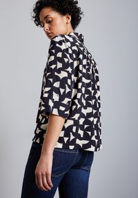 Camicia a maniche corte di colore blu navy con un motivo geometrico bianco, dotata di colletto e piega sul retro, abbinata a jeans scuri. Tessuto dalla texture liscia.