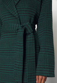 Cappotto a scacchi verde e nero con vita cintata, colletto a punta e tessuto testurizzato. Presenta maniche ampie e una finitura morbida.