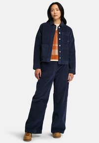 Marinblå kordjacka med framfickor och tryckknappar, i kombination med matchande vida byxor. Modellen bär en orange mönstrad tröja under.