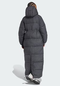 Duga crna zimska jakna s kapuljačom, s preklopnim dizajnom i elastičnim strukom. Zip zatvarač na dnu. Uparena s bijelim tenisicama.