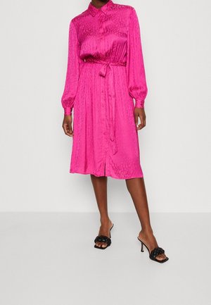 Mujer vistiendo un vestido midi de manga larga y color rosa brillante, con botones y cinturón anudado en la cintura, acompañado de sandalias de tacón alto negras con trenzado.