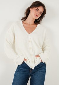 Cardigan alb pufos cu decolteu în V, nasturi mari și manșete cu nervuri, purtat cu blugi de culoare albastru închis. Textură moale vizibilă.