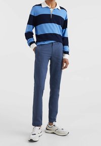 Persona vestida con una camiseta de rugby de rayas azules y largas mangas, pantalones azules y zapatillas blancas, de pie con una mano en el bolsillo sobre un fondo blanco.