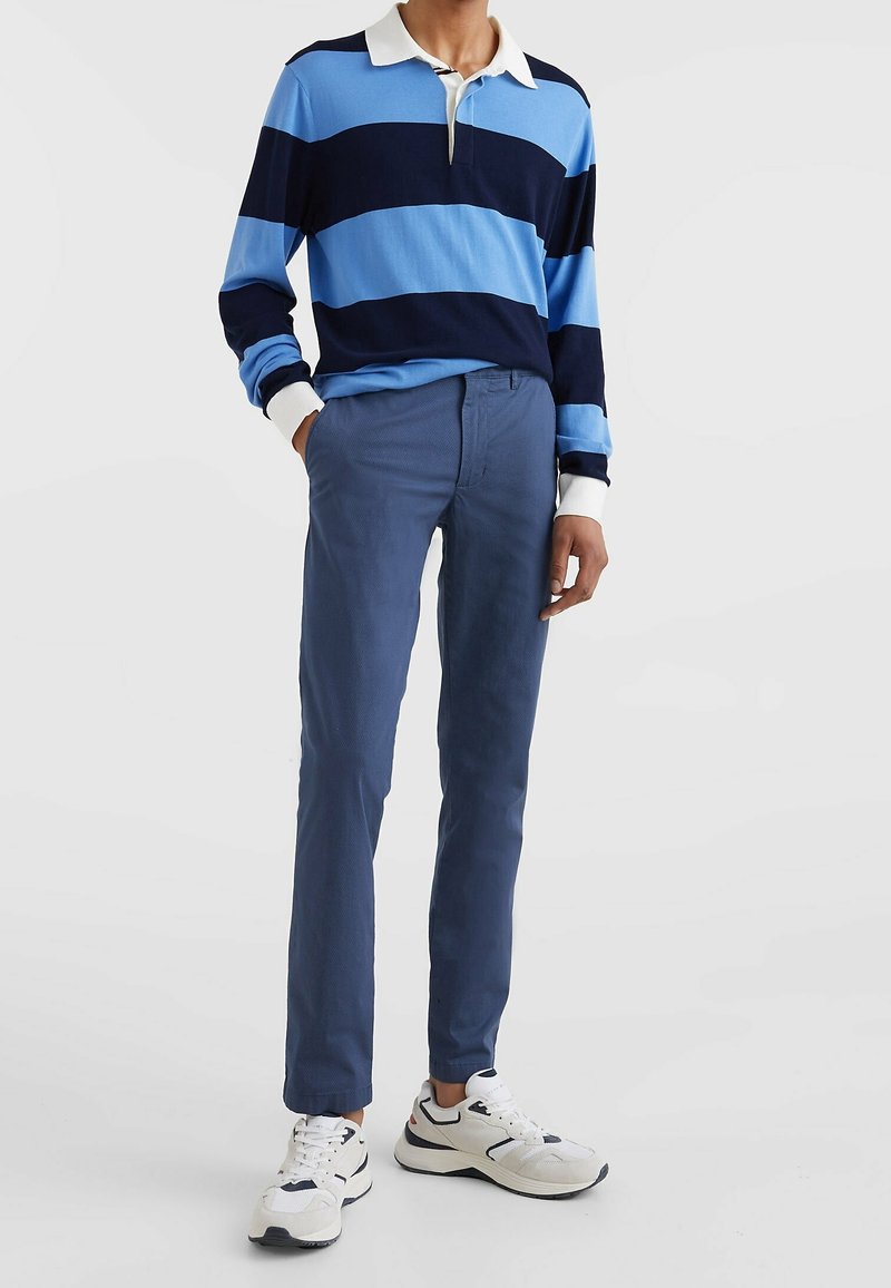 Persona vestida con una camiseta de rugby de rayas azules y largas mangas, pantalones azules y zapatillas blancas, de pie con una mano en el bolsillo sobre un fondo blanco.