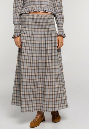 Mujer que lleva una falda gris de cuadros de talle alto y hasta el tobillo, con una blusa a juego de manga larga y zapatos planos marrones con cordones.