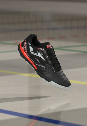 Svart innendørs sportssko med hvite og røde detaljer, skolissene er knyttet, svever over et gymsalgulv merket med fargede linjer.