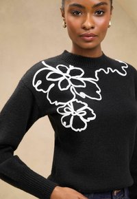 Maglione nero con maniche lunghe, caratterizzato da ricami floreali bianchi sulla spalla sinistra. Texture lavorata a maglia con scollatura a costine.