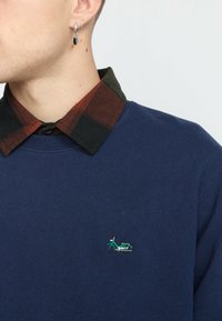 Marineblauwe sweatshirt met een textuurafwerking, voorzien van een groene fiets borduring op de borst en een geruite kraag in donkergroen en rood.