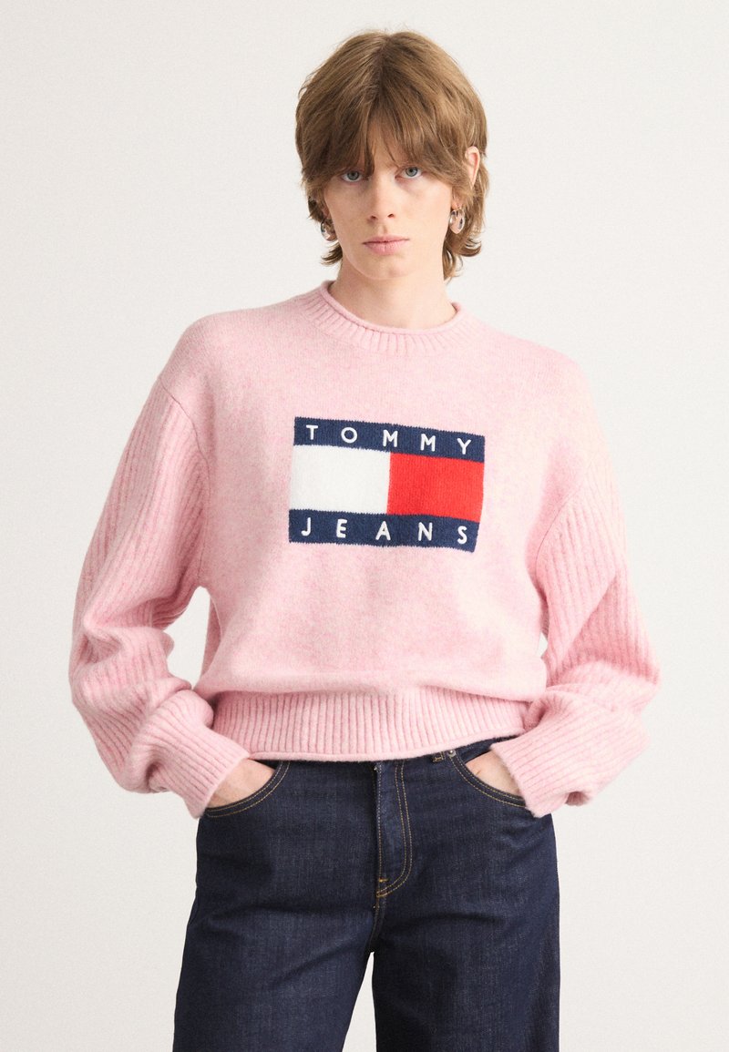 Rožinė megzta svają su räbotais rankogaliais ir apatine dalimi, turinti stačiakampį logotipo pleistrą tamsiai mėlynu, baltu ir raudonu su tekstu "TOMMY JEANS".
