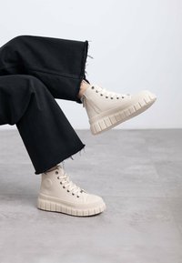 Zapatillas altas beige con una superficie texturizada y suela gruesa y estriada. Llevadas con unos jeans de pierna ancha negros deshilachados, ligeramente inclinados.