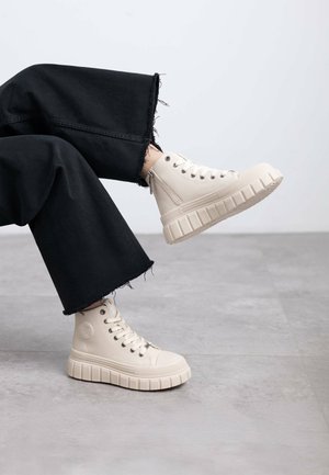 Zapatillas altas beige con una superficie texturizada y suela gruesa y estriada. Llevadas con unos jeans de pierna ancha negros deshilachados, ligeramente inclinados.