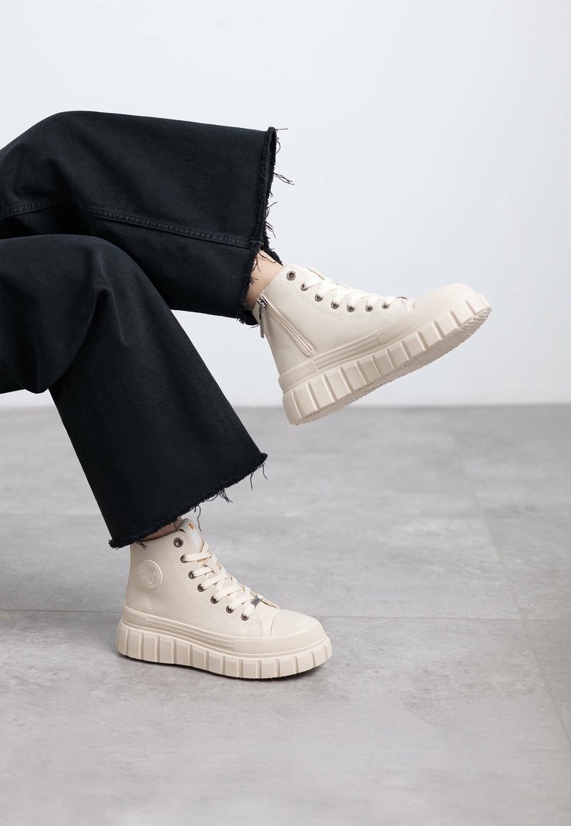 Zapatillas altas beige con una superficie texturizada y suela gruesa y estriada. Llevadas con unos jeans de pierna ancha negros deshilachados, ligeramente inclinados.