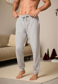 Pantaloni della tuta grigi con vita regolabile con coulisse, design a gamba affusolata e tessuto morbido. Presentano un dettaglio logo sul lato. Posizione a piedi nudi.