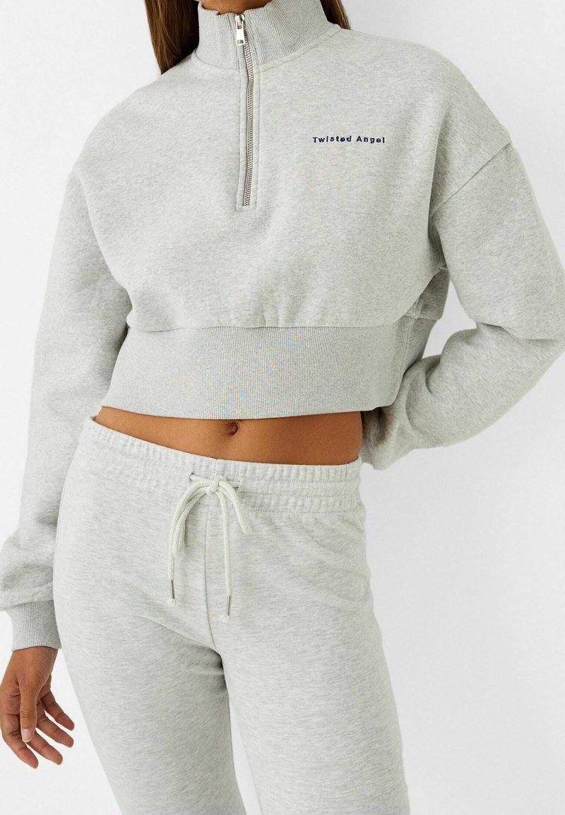Felpa grigia a maniche lunghe con zip a quarti e ricamo "Twisted Angel". Abbinata a pantaloni della tuta grigi con vita elastica.