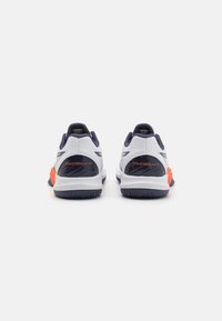 Vue arrière de chaussures de sport présentant une tige en mesh blanche avec des accents navy, un détail au talon orange et une semelle en caoutchouc texturée.