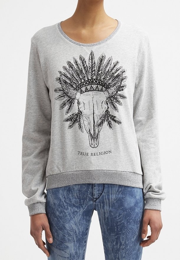 Personne portant un sweat-shirt gris clair avec un graphique de crâne de taureau noir et une coiffe de plumes ainsi que le texte "TRUE RELIGION", associé à un jean bleu à motifs.