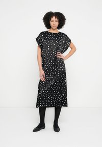 Vestido de cetim preto com bolinhas brancas, mangas curtas, comprimento até ao joelho, corte solto, com decote redondo e abertura lateral.