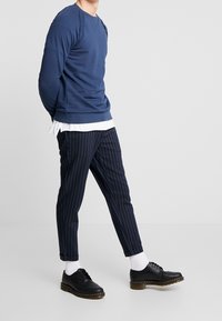 Marineblauwe sweatshirt met ribgebreide boorden, gecombineerd met marineblauwe broek met streepjes. Zwarte schoenen met ronde neusstukken, leren bovenwerk en rubberen zolen.