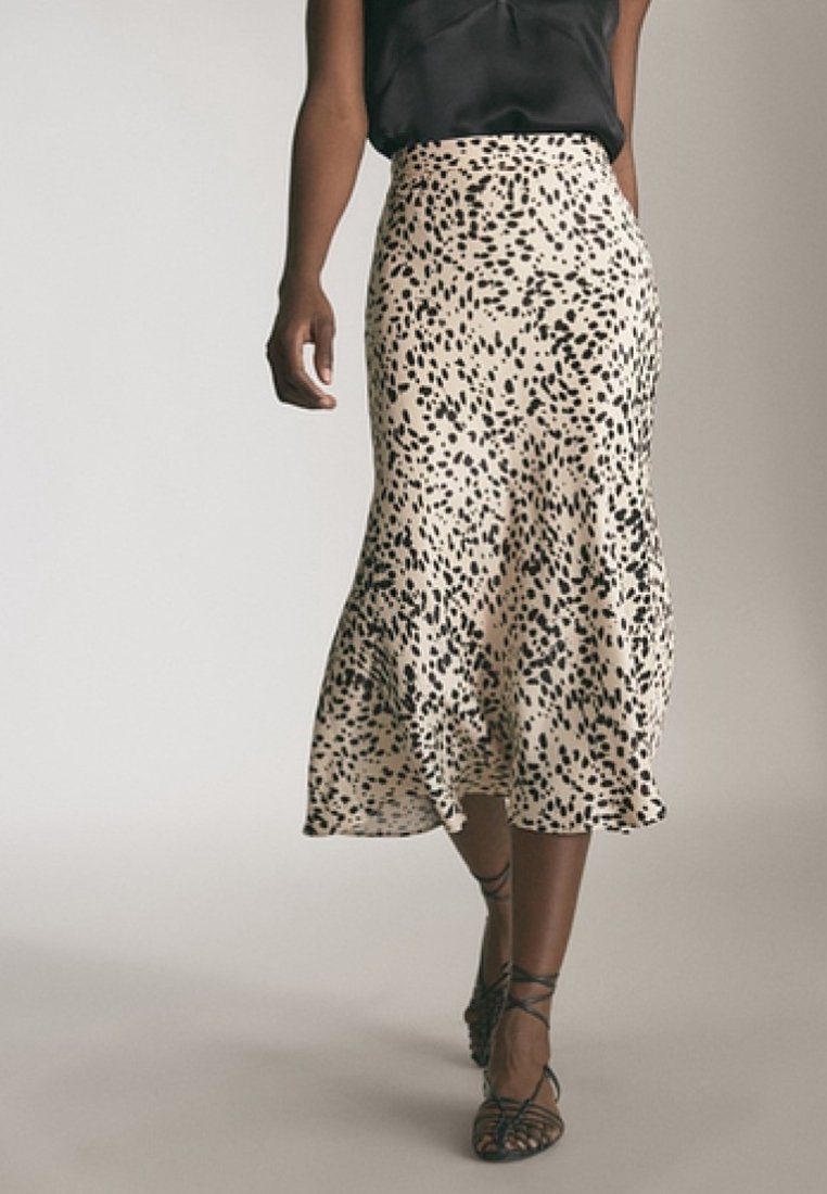 Femme portant un haut noir sans manches, une jupe midi beige avec des taches abstraites noires et des sandales noires à lanières marchant vers l'avant.