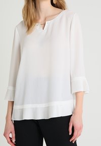 Valkoinen, pitkähihainen paita, joka on valmistettu sileästä kankaasta. Paidassa on V-aukko, jossa on kaksi nappia sekä laskostettu helma ja hihansuut.