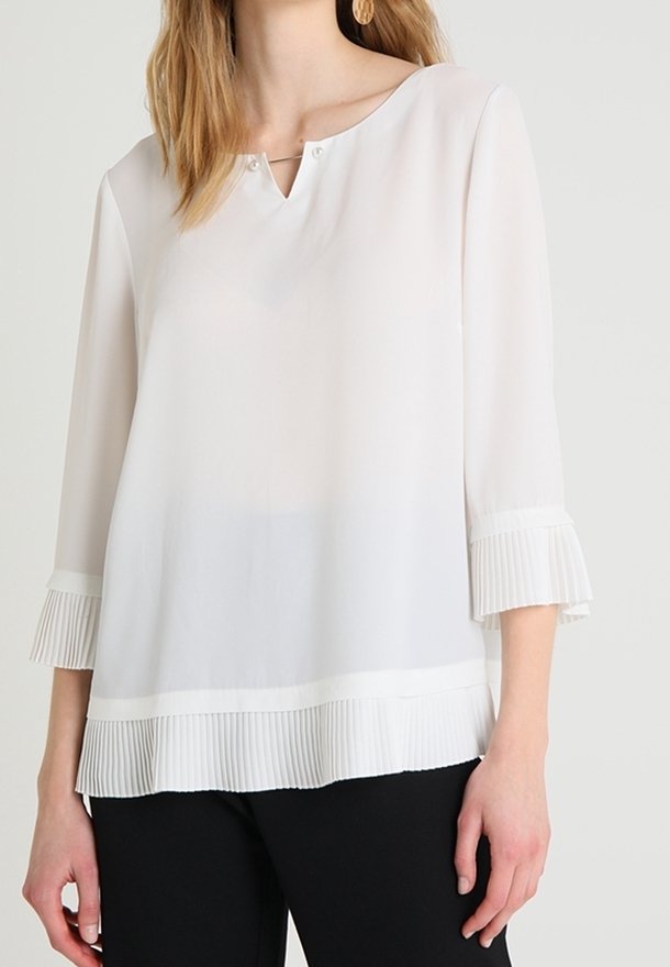 Valkoinen, pitkähihainen paita, joka on valmistettu sileästä kankaasta. Paidassa on V-aukko, jossa on kaksi nappia sekä laskostettu helma ja hihansuut.