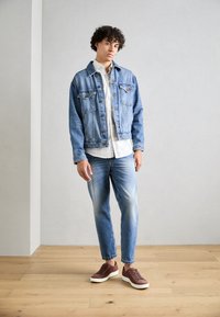 Veste en denim avec fermeture à boutons à l'avant, deux poches sur la poitrine, lavage bleu clair ; associée à une chemise blanche, un jean bleu clair et des baskets marron.