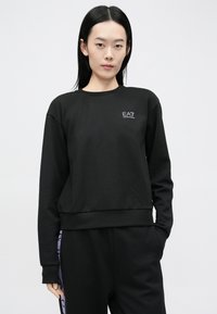 Žena s rovnými čiernymi vlasmi, oblečená v čiernej mikine EA7 a čiernych nohaviciach s fialovými bočnými pruhmi, stojaca pred bielym pozadím.