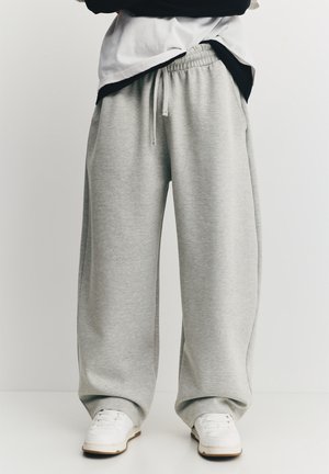 Pantalon de survêtement gris clair avec coupe décontractée, taille élastique et cordon de serrage. Assorti à des baskets blanches sur un fond uni.
