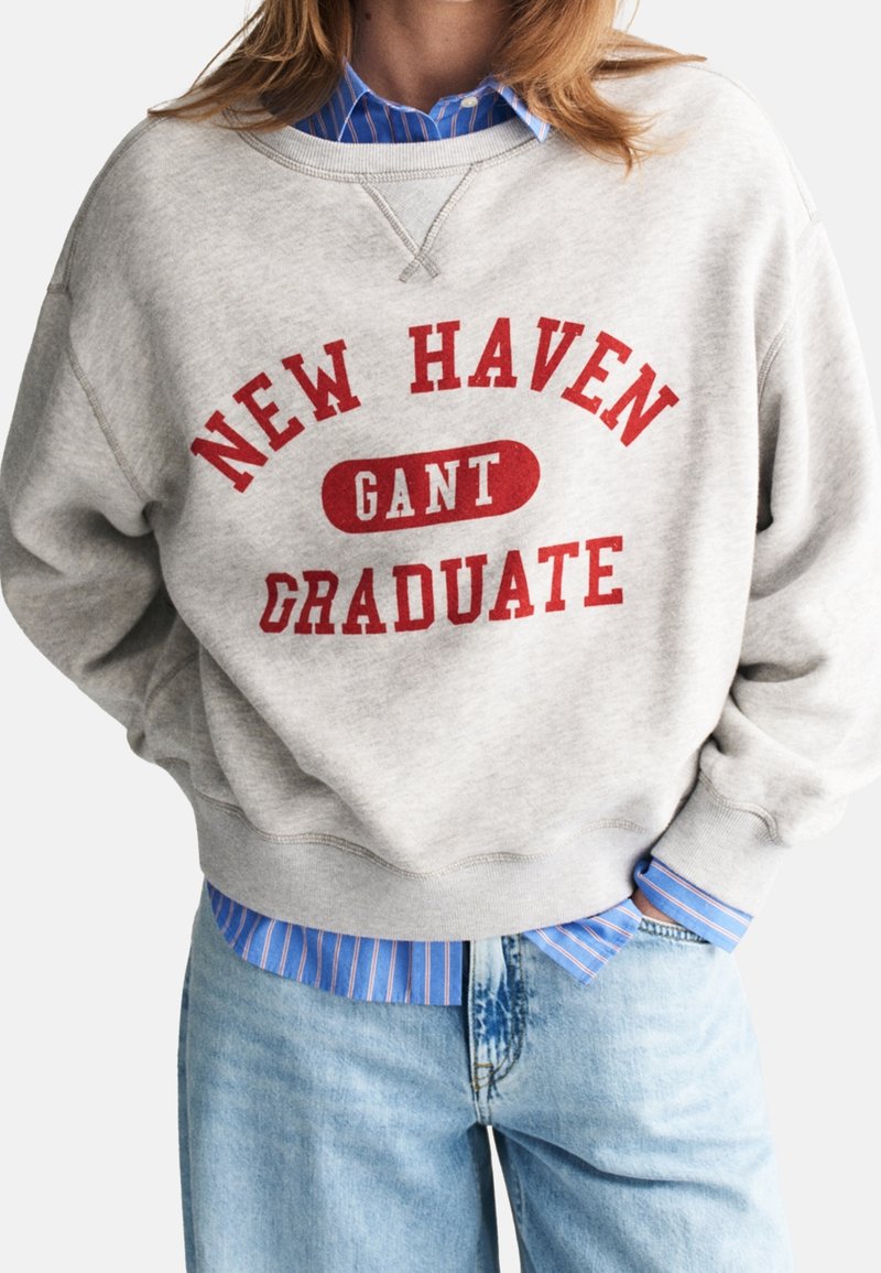 Personne portant un sweat-shirt gris clair avec un texte rouge "NEW HAVEN GANT GRADUATE", superposé sur une chemise bleue rayée boutonnée et un jean bleu clair.