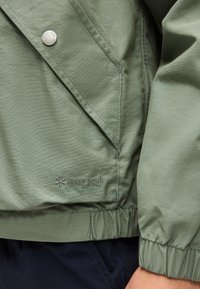 Manche et poche de veste verte avec bouton pression et logo brodé "snow peak" au-dessus de la ceinture élastique et pantalon foncé.