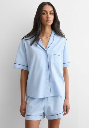 Frau trägt hellblaues kurzärmeliges Hemd mit Knöpfen und passende Shorts mit schwarzer Paspelierung, steht vor einem einfarbigen weißen Hintergrund.