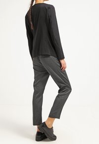 Kobieta w czarnym żakiecie, szarych dopasowanych spodniach i czarnych butach na platformie, stoi z lekko zgiętą prawą nogą na tle białym.