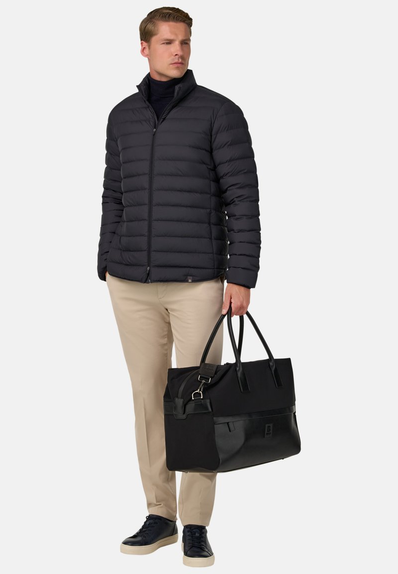 Svart vadderad jacka, beige byxor och marinblå sneakers. Håller i en svart och läder toteväska med dubbla handtag, som har ett blixtlåsstängning.