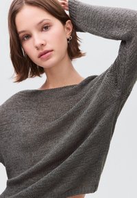 Junge Frau in einem lockeren grauen Strickpullover mit einem erhobenen Arm, kurzen braunen Haaren und sternförmigen Ohrringen vor einem schlichten Hintergrund.