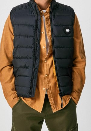 Mężczyzna w ciemnej pikowanej kamizelce na musztardowej koszuli zapinanej na guziki i oliwkowych spodniach, ręce w kieszeniach, z logo "PEPE JEANS LDN" na piersi.
