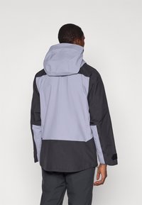 Homem a usar uma jaqueta impermeável bicolor com capuz, visto de costas contra um fundo branco simples.