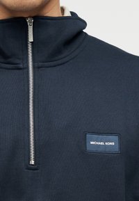 Μπλε σκούρο πουλόβερ με ψηλό γιακά, ασημένιο φερμουάρ και ορθογώνια ετικέτα Michael Kors στο στήθος, με υφή υφάσματος.
