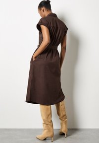 Robe marron, longueur genou, avec manches courtes, poches latérales et taille ceinturée ; accompagnée de bottes beige de hauteur genou à bout pointu.