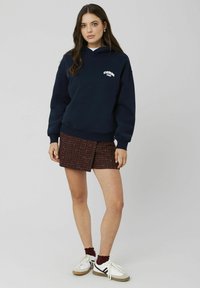 Hoodie bleu marine avec un logo sur la poitrine, associé à une jupe texturée courte bourgogne et des baskets blanches avec des accents noirs.
