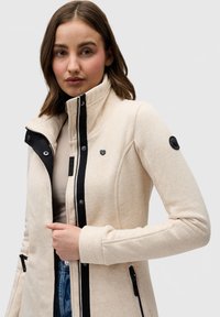Béžová bunda s vysokým golierom, kontrastnými čiernymi zipsami a bočnými vreckami. Mäkká štruktúra s hladkým povrchom a logom na rukáve.