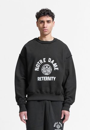 Jonge man met krullend haar die een zwart sweatshirt en broek draagt, beide bedrukt met "NOTRE DAME" en een rond embleem.