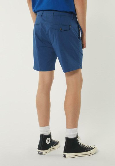 Pantaloni blu realizzati in tessuto leggero, con tasche posteriori con chiusura a bottone. Indossati con sneakers alte nere e calzini bianchi.
