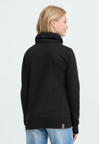 Felpa nera con colletto alto, polsini e orlo a coste. Etichetta marrone chiaro ben visibile sul fondo dell'orlo. Materiale morbido e testurizzato.