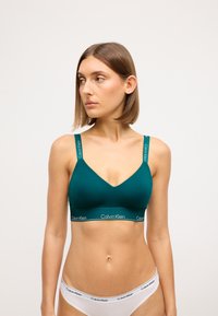 T türkiz bralette V-kivágással, állítható pántokkal és "Calvin Klein" feliratos elasztikus szalaggal. Sima textúra, minimalista stílus.